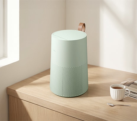 A mint-green LG air purifier with a brown leather strap sits on a wooden desk next to a white cup of tea in a bright, sunlit room.	
