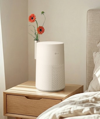 A white LG air purifier on a wooden bedside table, functioning as a decor piece with a silver vase holding two orange flowers in a sunny bedroom.	