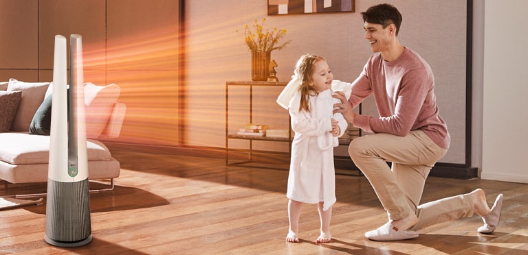Father kneeling on the floor, wrapping his daughter in a robe, while LG heater emits warm air in the living room.