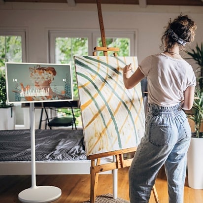 A woman paints a picture. There is standbyME next to it and music is playing on the screen.