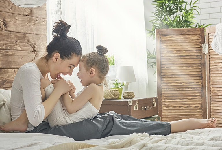 Woman and child sit on a bed in a sunlit room with wooden decor, plants, and natural light streaming through a window.