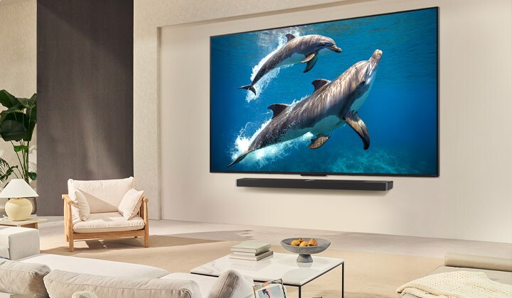A family sits on a couch facing a LG QNED TV mounted on a wall with a little girl pointing to a screen depicting two dolphins.