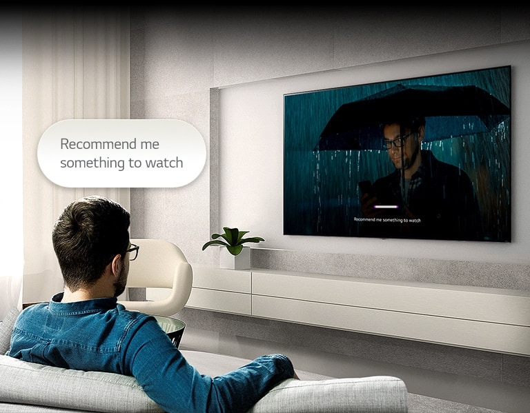 A man is sitting on a sofa facing a TV on the opposite side. A speech bubble that reads 'Recommend me something to watch' is floating above his head. Inside the screen, a video of a man wearing an umbrella is playing with a UI that is voice-recognized.