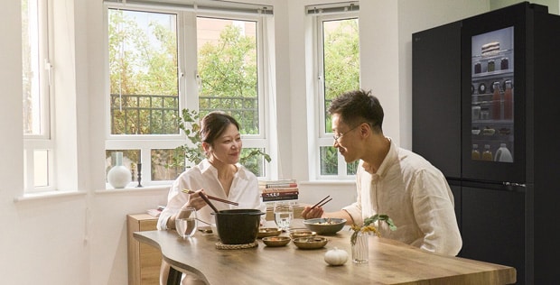 Egy pár ül az étkezőasztalnál, és esznek, mellettük egy LG Side-by-side InstaView hűtőszekrény áll.