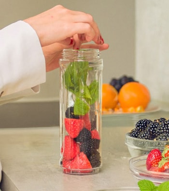 Egy nő epret, szedret és fűszereket tesz egy hosszúkás üveg edénybe, körülötte üvegtálak találhatók tele a mocktail hozzávalóival.