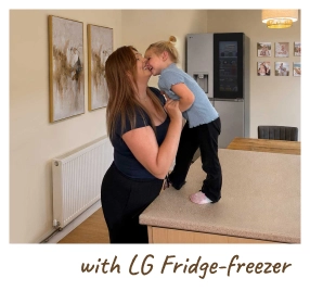 Egy édesanya a karjában tartja a babáját egy modern konyhában, egy nyitott hűtőszekrény mellett állva. [with LG Fridge-freezer]