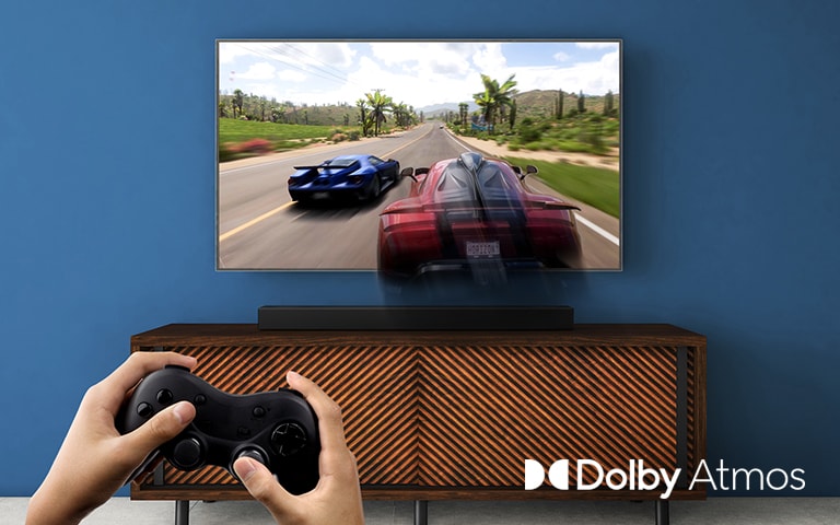 Egy LG TV a falra van felszerelve, és egy versenyjáték látható rajta. Az LG hangprojektor egy barna polcra van helyezve, közvetlenül az LG TV alá. Egy férfi egy botkormányt tart.