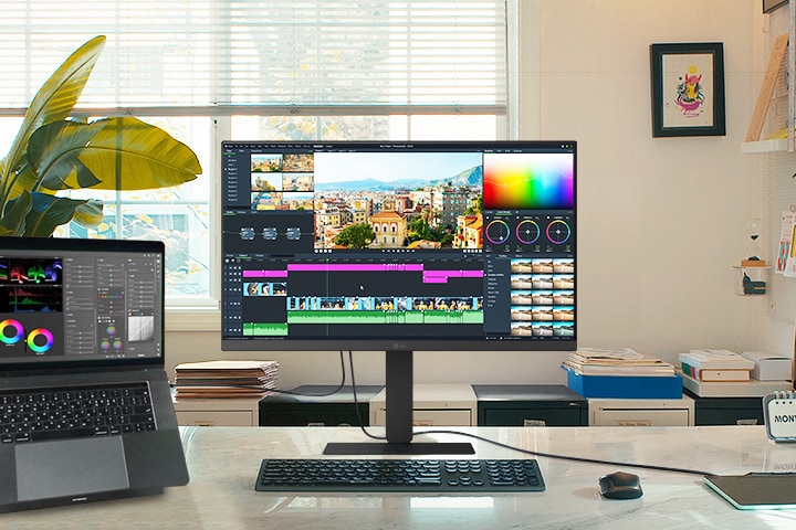 Egy kreatív munkaterület egy LG monitorral és egy csatlakoztatott laptoppal, mindkettő egy videószerkesztő felületet jelenít meg színkorrekciós eszközökkel és idővonallal.