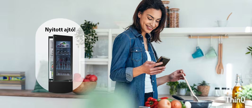A képen egy nő főzés közben értesítést kap a mobiltelefonjára az LG ThinQ applikációból, a nyitva felejtett hűtő ajtó miatt.