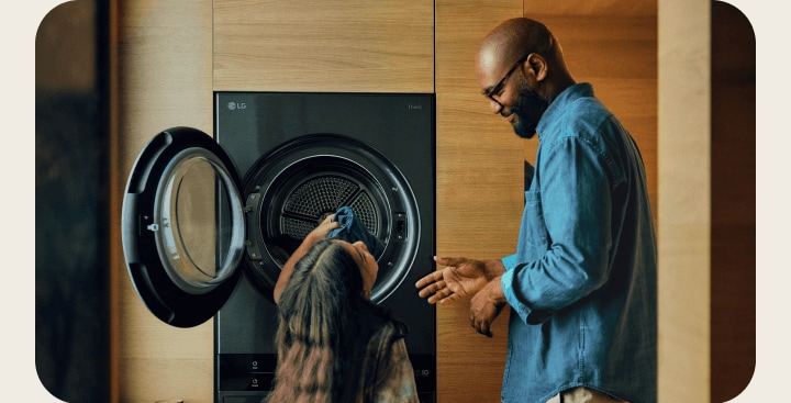 A father and daughter taking laundry out of an open LG WashTower AI, next to an LG Robot Vacuum cleaning the floor beneath a dining table.