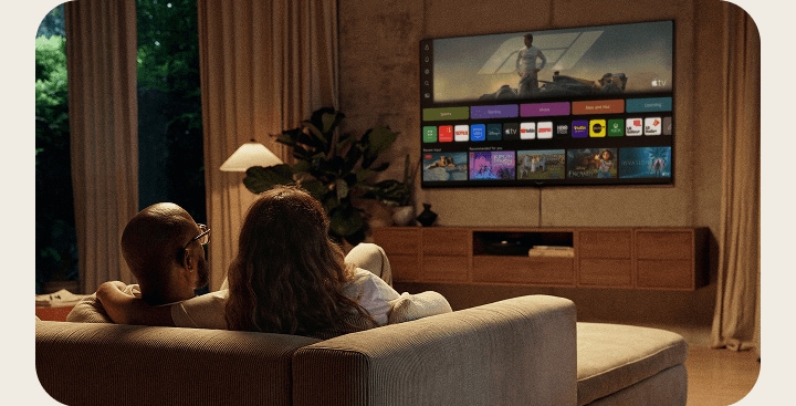 An LG xboom AI speaker resting on a side table, next to a couple relaxing on a sofa watching LG Channels on their LG AI TV.