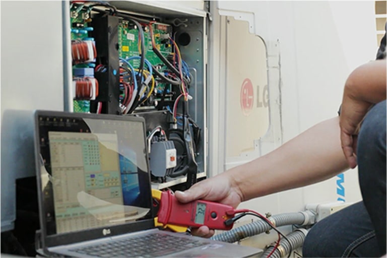 Seseorang menggunakan alat persegi panjang merah yang terhubung ke laptop, mendiagnosis unit luar ruangan LG yang terbuka, kabel internalnya terbuka sepenuhnya.