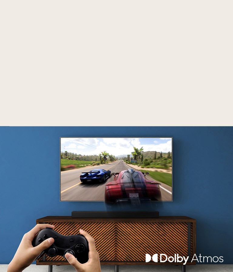 TV LG berada di dinding, menampilkan permainan balap. LG Sound Bar diletakkan di rak coklat, tepat di bawah TV LG. Seorang pria memegang joy stick.