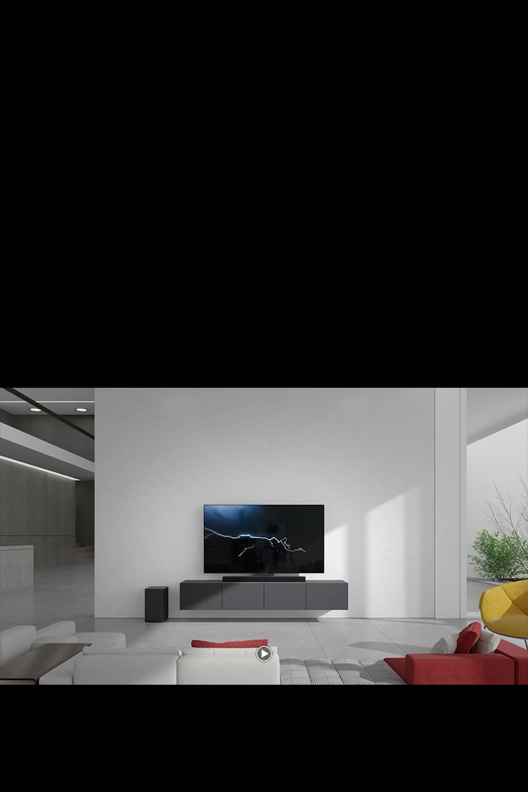 Sound bar ditempatkan pada kabinet abu-abu dengan TV di ruang tamu. Subwoofer nirkabel hitam diletakkan di lantai di sisi kiri dan sinar matahari masuk dari sisi kanan gambar. Sofa panjang berwarna putih dan merah ditempatkan menghadap TV dan sound bar.