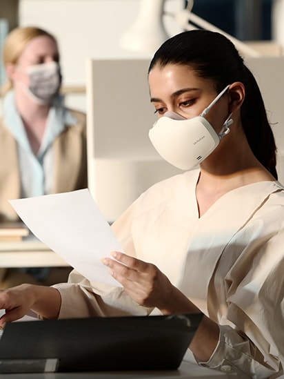 Seorang perempuan sedang memperhatikan dokumen didalam sebuah kantor, dengan LG PuriCare™ Wearable Air Purifier putih yang menyala