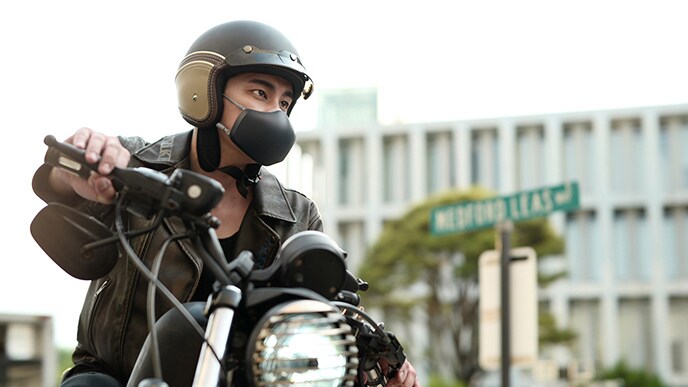 Seorang laki-laki Asia sedang mengendarai sepeda motor dengan menggunakan LG PuriCare™ Wearable Air Purifier warna hitam yang menyala