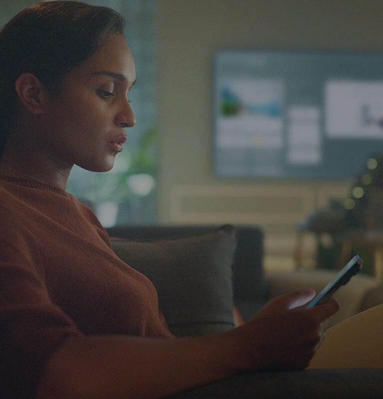 Image shows a woman looking into her phone in her livingroom sofa while TV is showing homeboard at the background.