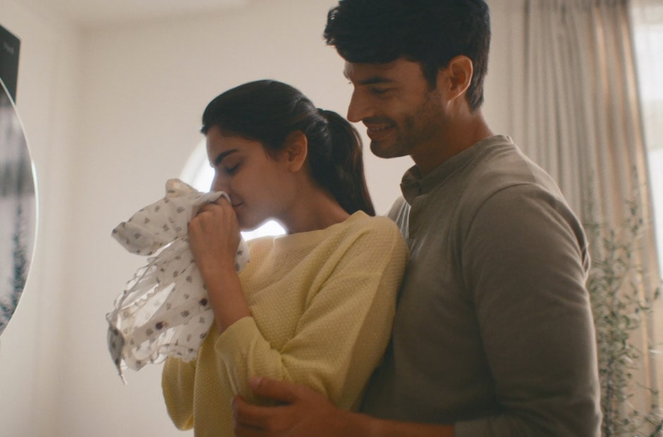 Image shows a man hugging a woman from behind as she smells a piece of freshly-washed baby clothing.