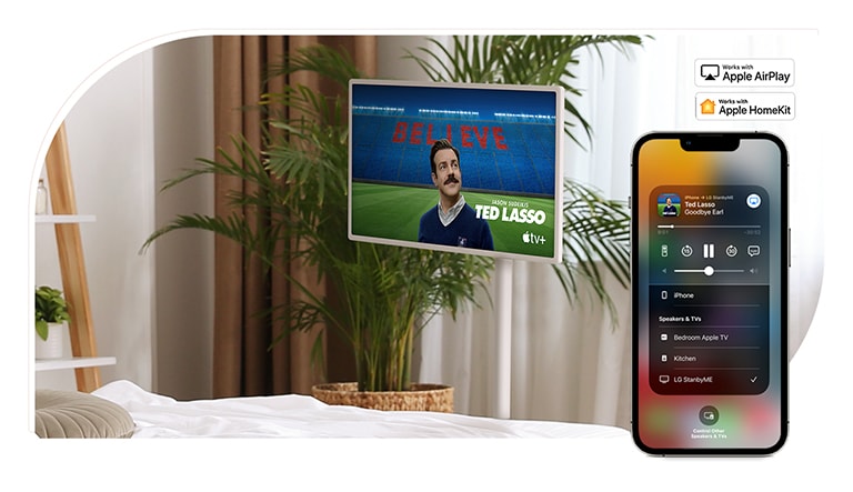 Sebuah TV ditempatkan di kamar tidur yang nyaman dan layarnya menampilkan acara TV – TED LASSO. Terdapat perangkat seluler pada gambar yang sama yang menampilkan AirPlay UI di layarnya.​ Ada logo Apple AirPlay dan logo Apple HomeKit ditempatkan di pojok kanan atas gambar.​