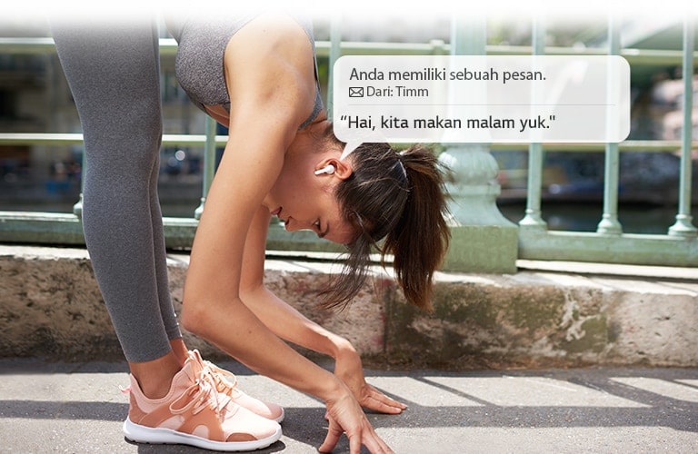 Seorang wanita sedang meregangkan badan dan membungkuk menyentuh tanah sambil mendengarkan pesan yang dibacakan earbud untuknya.