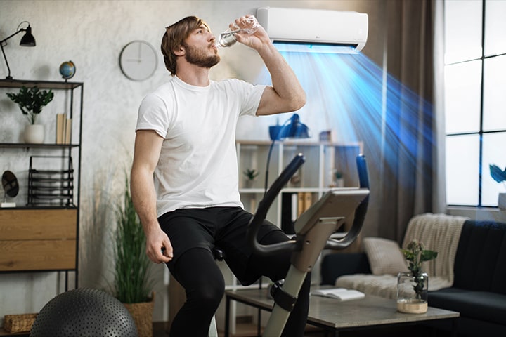 This is a video of the air conditioner wind coming out behind a man sitting on an exercise machine drinking water.