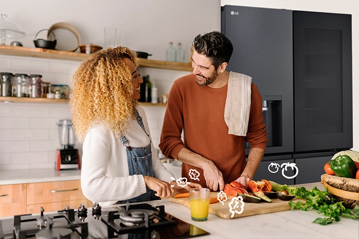 The smiling couple is cooking in front of the refrigerator.