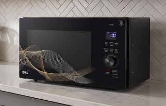 A silver countertop microwave sits in a modern kitchen with a gray stone backsplash, a sink, and a built-in oven.