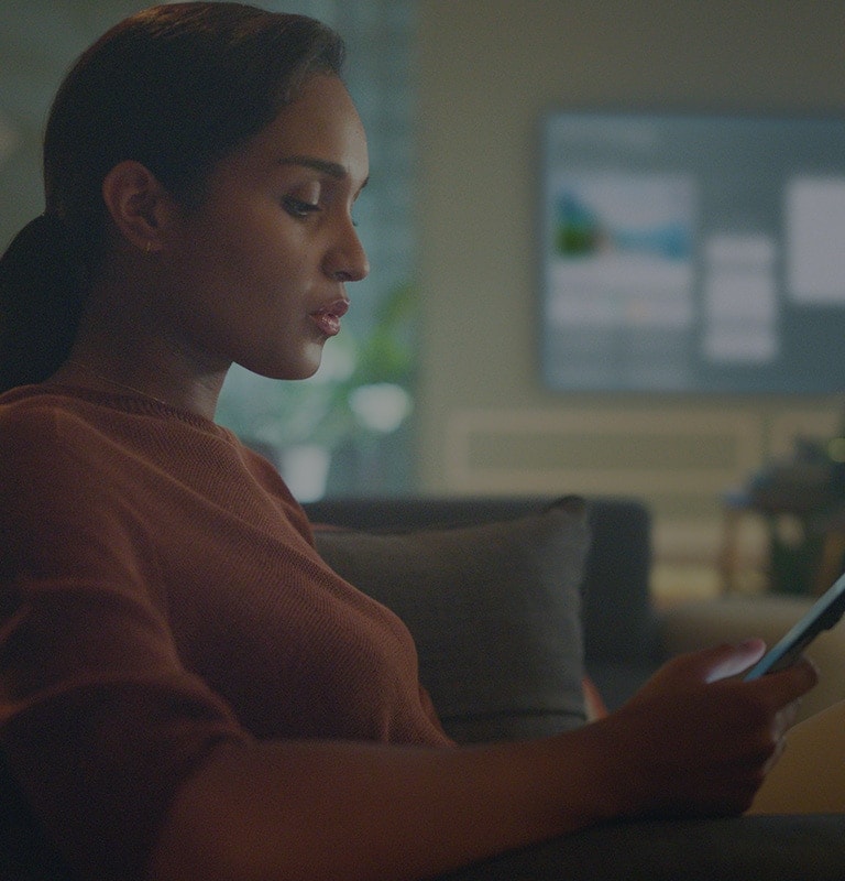 Image shows a woman looking into her phone in her living room sofa while TV is showing homeboard at the background.