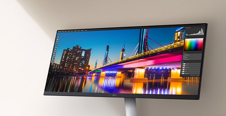 LG ultra-wide monitor displaying a vivid nighttime cityscape with a brightly lit bridge and reflection, shown within a photo editing software interface highlighting colour and contrast tools.