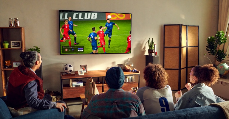 A man and woman sat behind a coffee table in front of a wall mounted TV showing a football match.