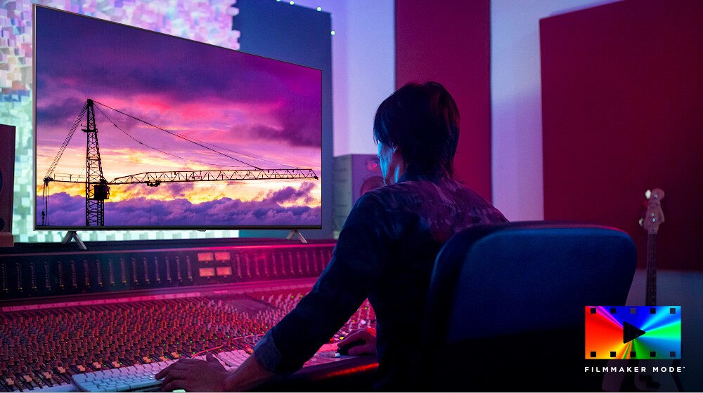 A movie director is looking at a big TV monitor, editing something. The TV screen shows a tower crane in purple sky. FILMMAKER Mode logo is placed on bottom right corner.