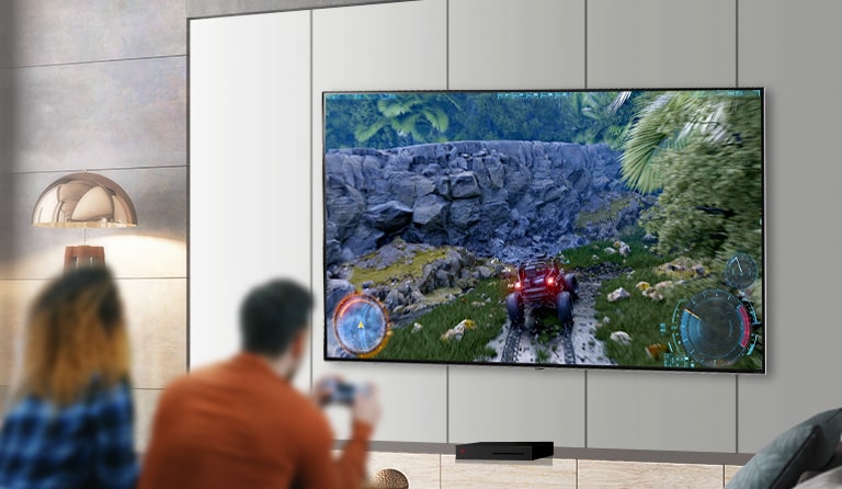 Rear view of a man and woman sat side-by-side in front of a large wall-mounted TV. The man is holding a gaming controller with a driving game on screen.