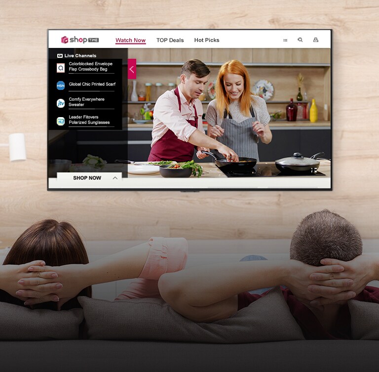 Couple sitting on a couch watching a TV displaying a man and a woman cooking in a kitchen and program information to shop.