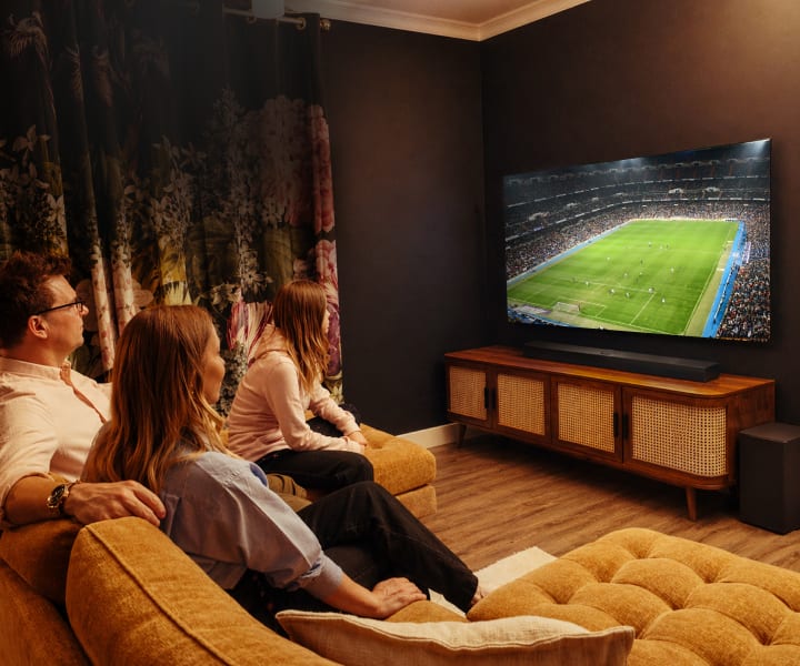 Una famiglia di tre persone siede insieme su un divano in un salotto accogliente e debolmente illuminato, guardando una partita di calcio su un grande TV OLED LG. La stanza è caratterizzata da pareti scure, tende floreali, un mobile media in legno e altoparlanti, che creano un'atmosfera calda e intima.