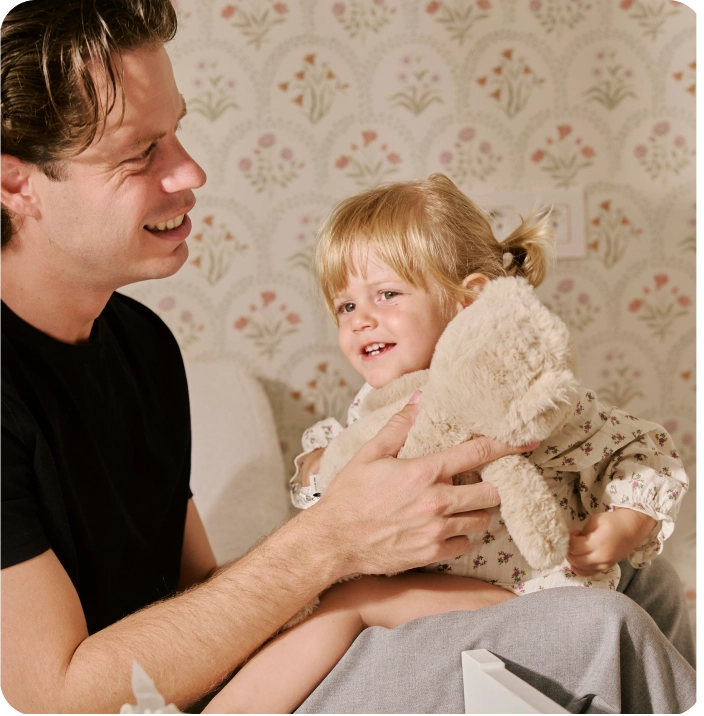 Un padre sorridente con una maglietta nera tiene in braccio la sua bambina bionda mentre lei gioca con un peluche.