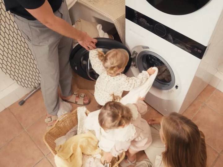 Vista dall’alto di una famiglia che fa il bucato insieme in una lavanderia piastrellata: i gemelli, ancora piccoli, tirano fuori i vestiti da una lavatrice bianca con asciugatrice sovrapposta, mentre i genitori li aiutano accanto a un cesto della biancheria intrecciato.