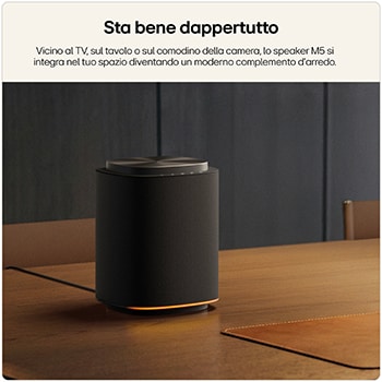 Speaker smart nero cilindrico su un tavolo in legno in un ambiente moderno; sullo sfondo una parete scura e una sedia.