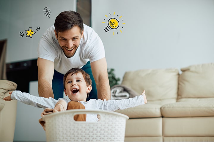 Un padre spinge il figlio nel cesto della biancheria. Intorno a loro ci sono disegni di lampadine, fiori e foglie che simboleggiano il risparmio energetico e la natura.