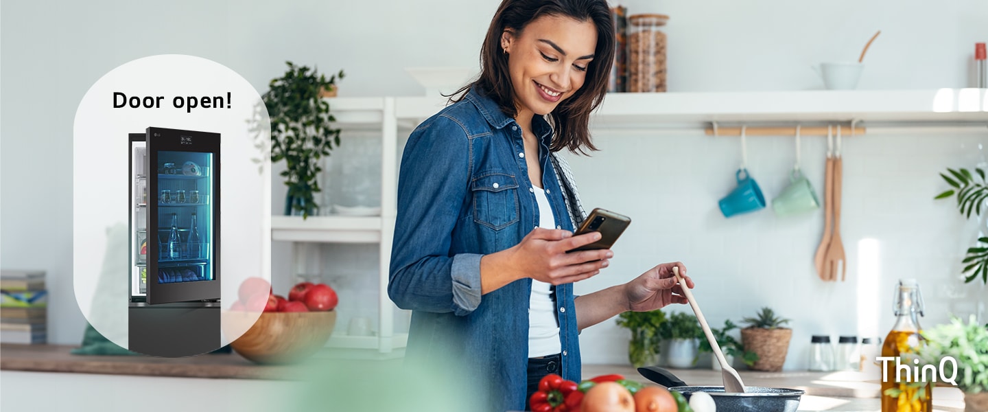 Immagine di una donna che cucina mentre guarda lo smartphone con dietro un frigorifero con la porta aperta.