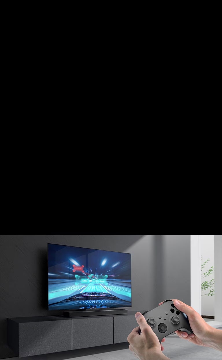 La barra audio viene posizionata sul mobile e la scena del gioco di corse viene mostrata sulla TV collegata alla soundbar. Una console di gioco si trova in basso a destra nell'immagine ed è tenuta da due mani.