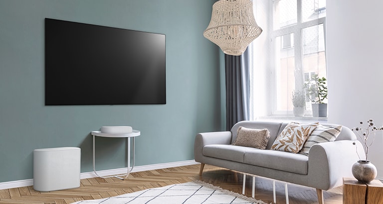 Una soundbar è collocata su un piccolo tavolino, mentre un subwoofer si trova sulla sinistra e un TV sopra la soundbar. L’intera stanza è bianca.