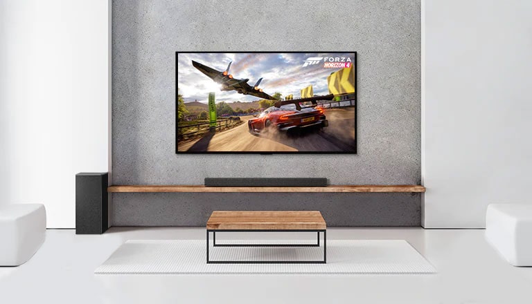 Un set di 2 altoparlanti posteriori, subwoofer e soundbar e un TV in  un salotto bianco. Sullo schermo del TV vediamo un’auto in corsa e una navicella spaziale.