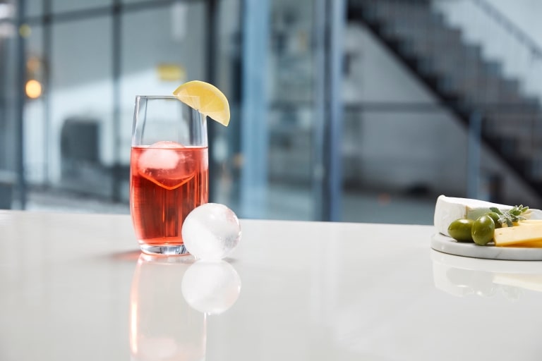 Un primo piano di una bevanda rinfrescante con ghiaccio e una fetta di limone, posta su un piano di lavoro bianco con un piatto di formaggio e olive sullo sfondo.