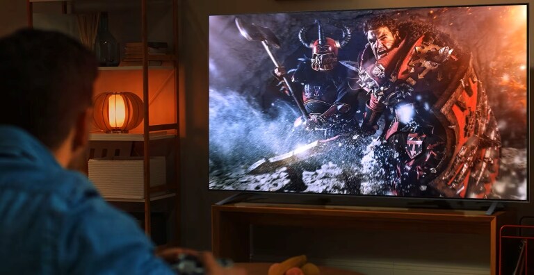 Una persona gioca a un videogioco su una TV LG. Sullo schermo, due guerrieri in armatura combattono in una scena intensa, mentre una lampada illumina la stanza con una luce calda.