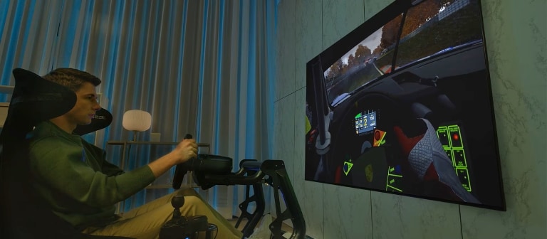 Una persona gioca a un simulatore di guida con volante e postazione realistica. Sul grande TV LG di fronte a lui appare una visuale interna dell'auto in corsa su un circuito, creando un'esperienza coinvolgente.