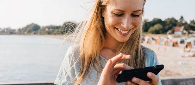 Una donna che sorride mentre usa il suo smartphone in spiaggia.