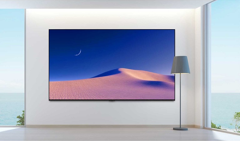 Un luminoso soggiorno moderno con un grande schermo che mostra una duna di sabbia e una luna crescente, accanto a una lampada da terra e ampie finestre con vista sull'oceano.