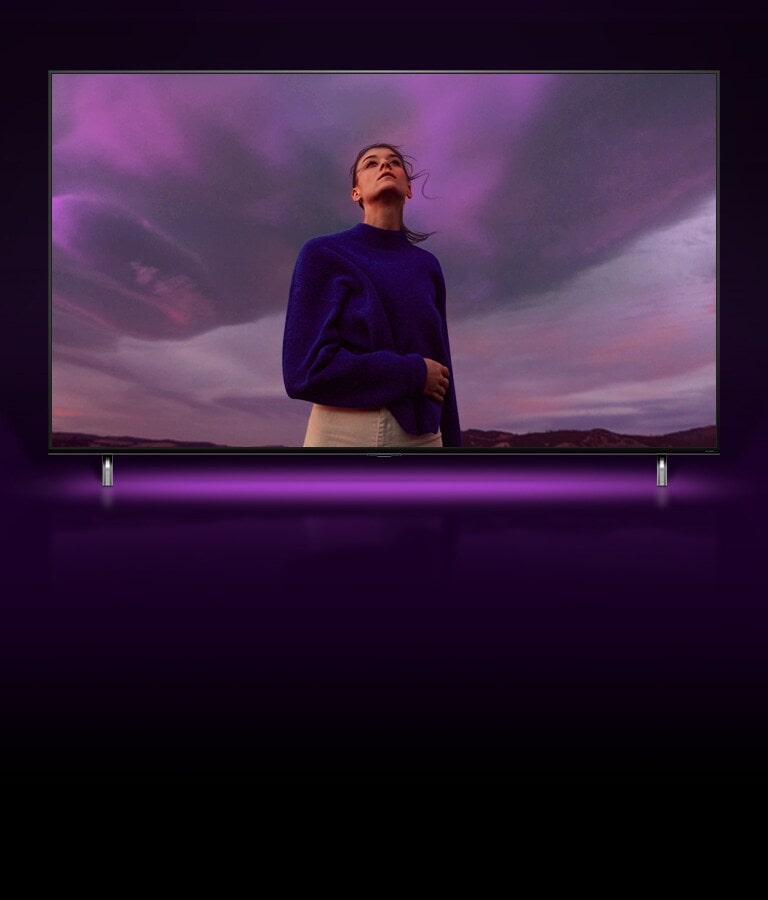 Una donna in piedi davanti a un cielo viola, poi l’inquadratura si allarga per mostrare la stessa donna sullo schermo TV QNED.