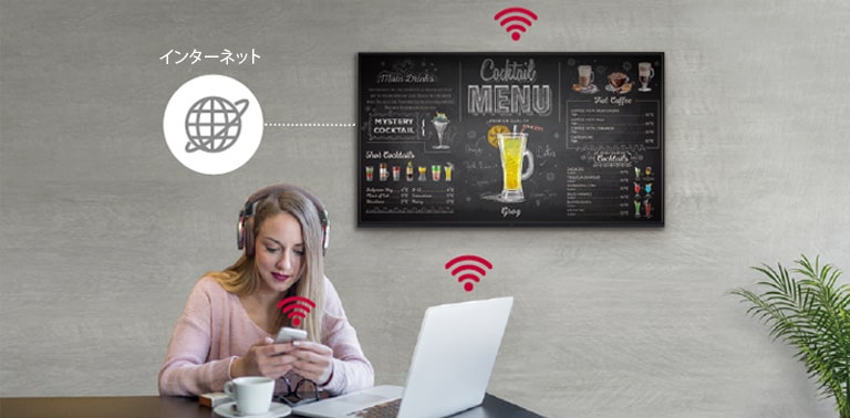 UR3Jシリーズが壁に設置されていて、女性がノートパソコンとスマートフォンを使用している。この画像は、パソコンや女性が手にしているスマートフォンと、サイネージをワイヤレス接続できることを示している。