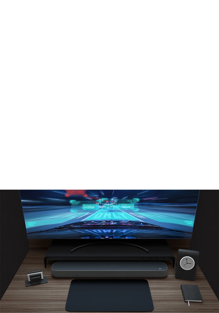 Sound Bar、湾曲したモニター、ノート、小さい時計が木製の机に置かれている。ゲーマー向けの機能を紹介するため、モニターにはレーシングゲームが映っている。